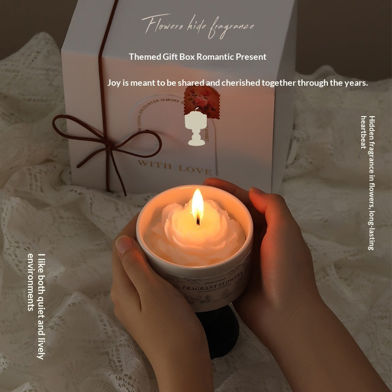 A person holding a burning rose-shaped flower candle in a ceramic chalice container, with an elegant white gift box and lace background.
