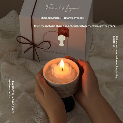 A person holding a burning rose-shaped flower candle in a ceramic chalice container, with an elegant white gift box and lace background.