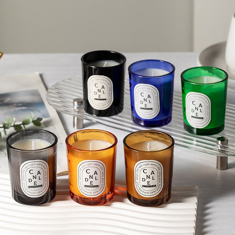 Six Delphy Candle scented candles in black, blue, green, smoke, orange and amber glass jars, neatly displayed on a clear glass tray for elegant home decor.