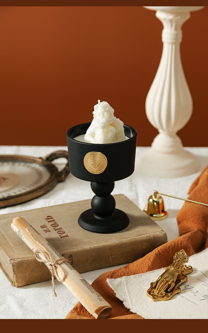 A white angel sculpture-shaped soy wax candle inside a matte black porcelain goblet with a gold seal, placed on a vintage book.